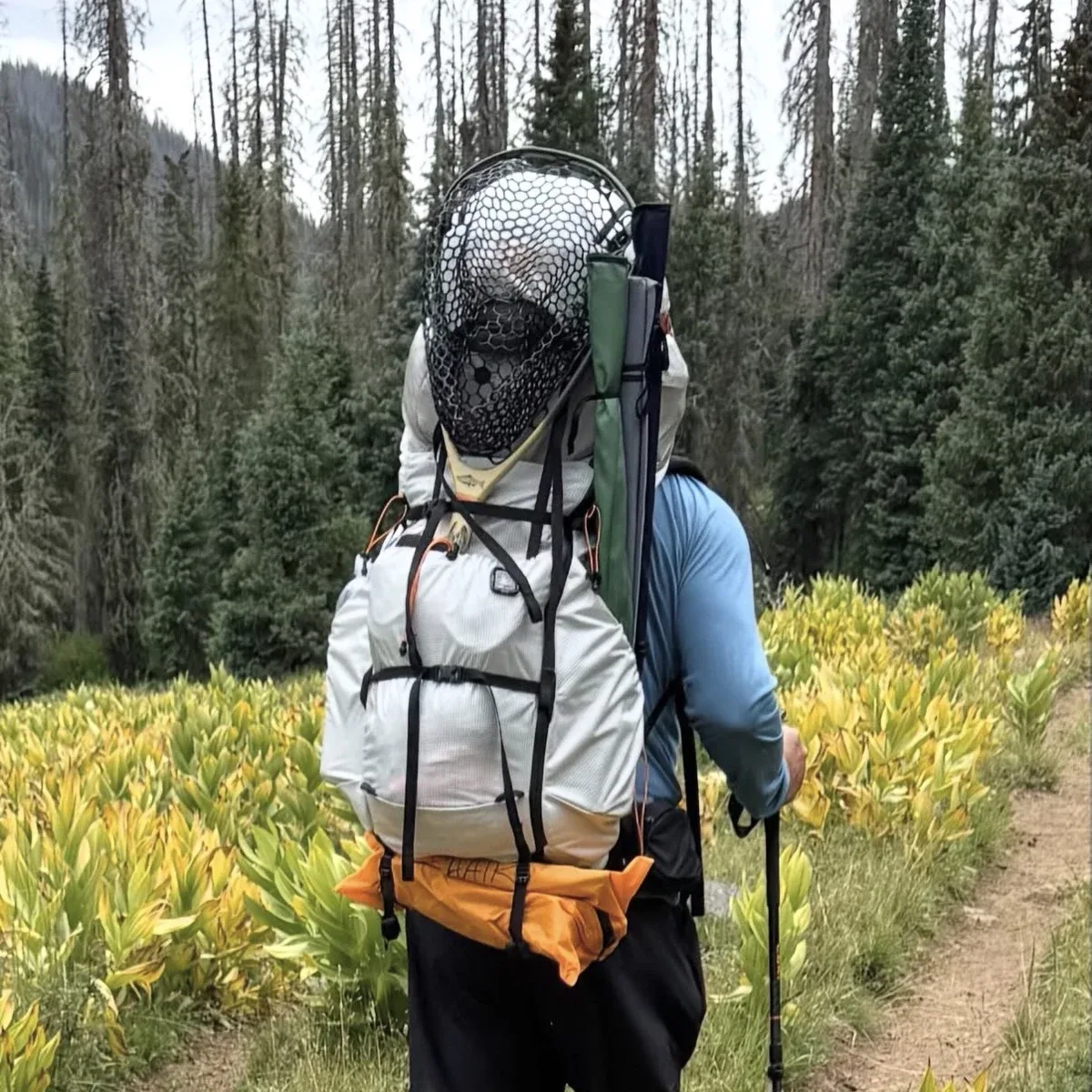 Kevin from behind on a forested backcountry trail, fly fishing net and rod tube strapped to an ultralight backpacking pack.